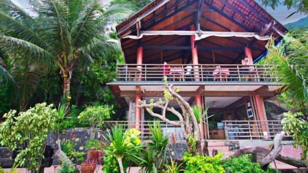 A two-story building with a wooden roof and balcony, surrounded by lush greenery and palm trees. - Planet Dive (Mabini)