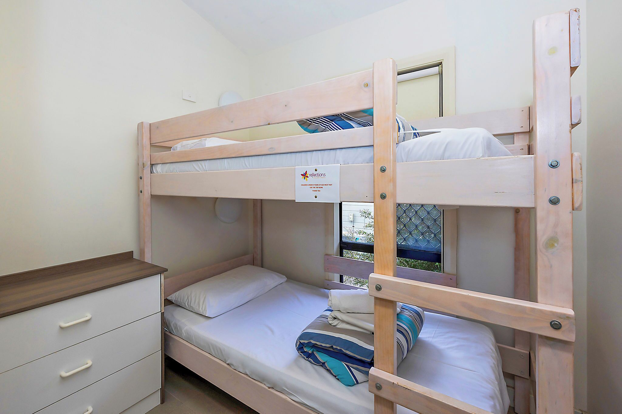 Una cama litera con estructura de madera, una mesita de noche pequeña y un módulo de cajones.