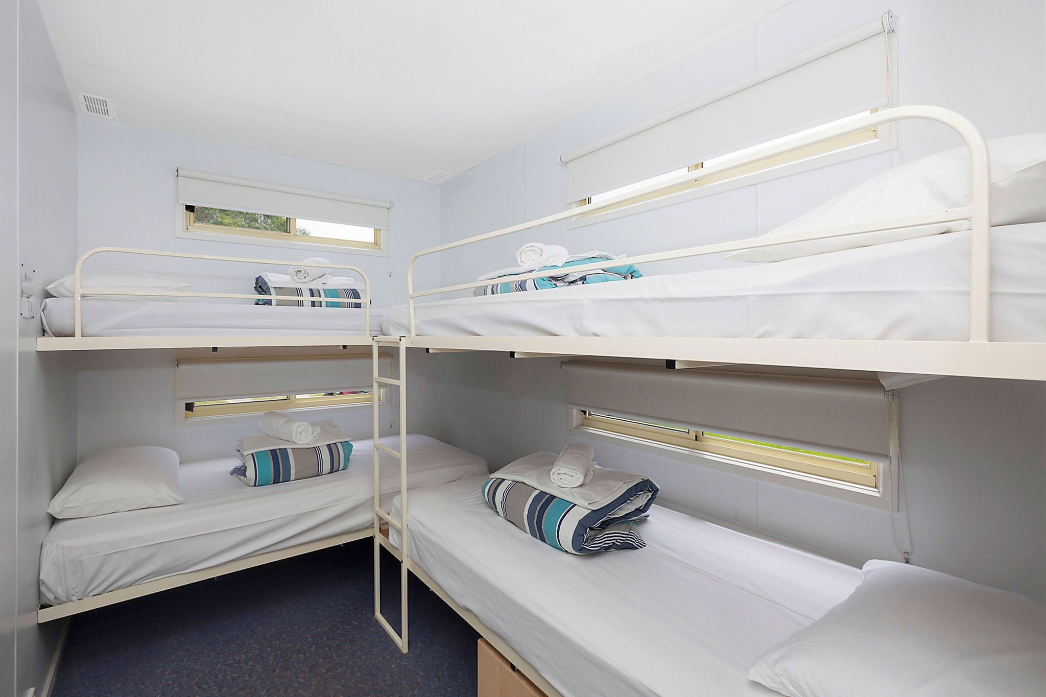 A bunk bed room with three beds, each with a pillow and a blanket. There is a window above the beds, and a ladder is attached to the wall.