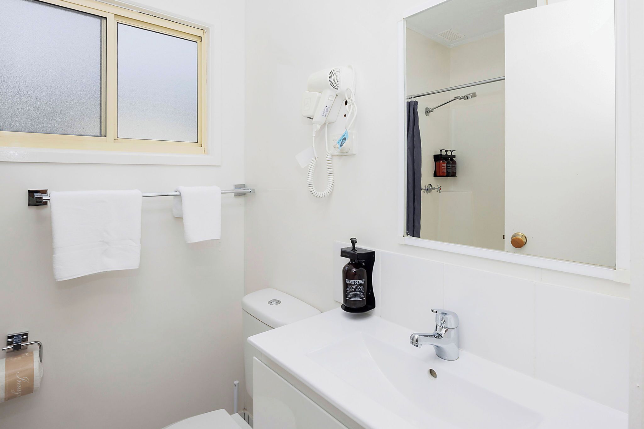 Une salle de bain avec un lavabo blanc, un sèche-cheveux, une bouteille de shampoing et un porte-serviettes.