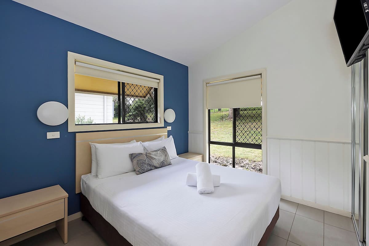 A hotel room with a large bed, a nightstand, a window with a view of greenery, and a TV mounted on the wall.