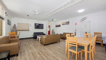 Amplia sala de estar con un televisor de pantalla plana grande, diversas opciones de asientos que incluyen sofás y sillones, una mesa de comedor de madera y un ventilador de techo.