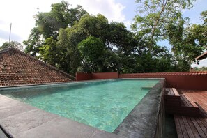 A rectangular swimming pool with clear water, surrounded by a wooden deck and a tiled roof. - Petit Hotel Bali (Ubud)