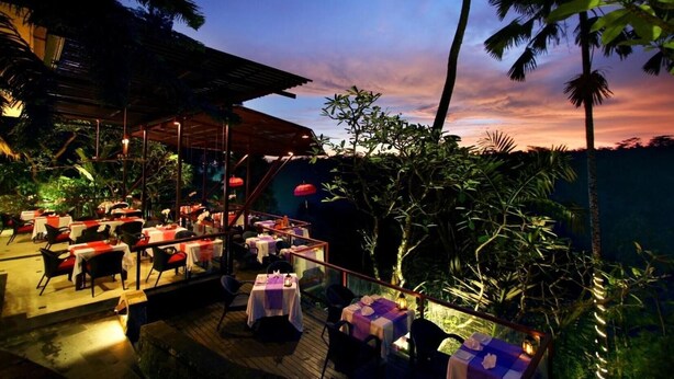 An outdoor restaurant with tables and chairs, surrounded by tropical vegetation and palm trees, under a sunset sky. - Petit Hotel Bali (Ubud)