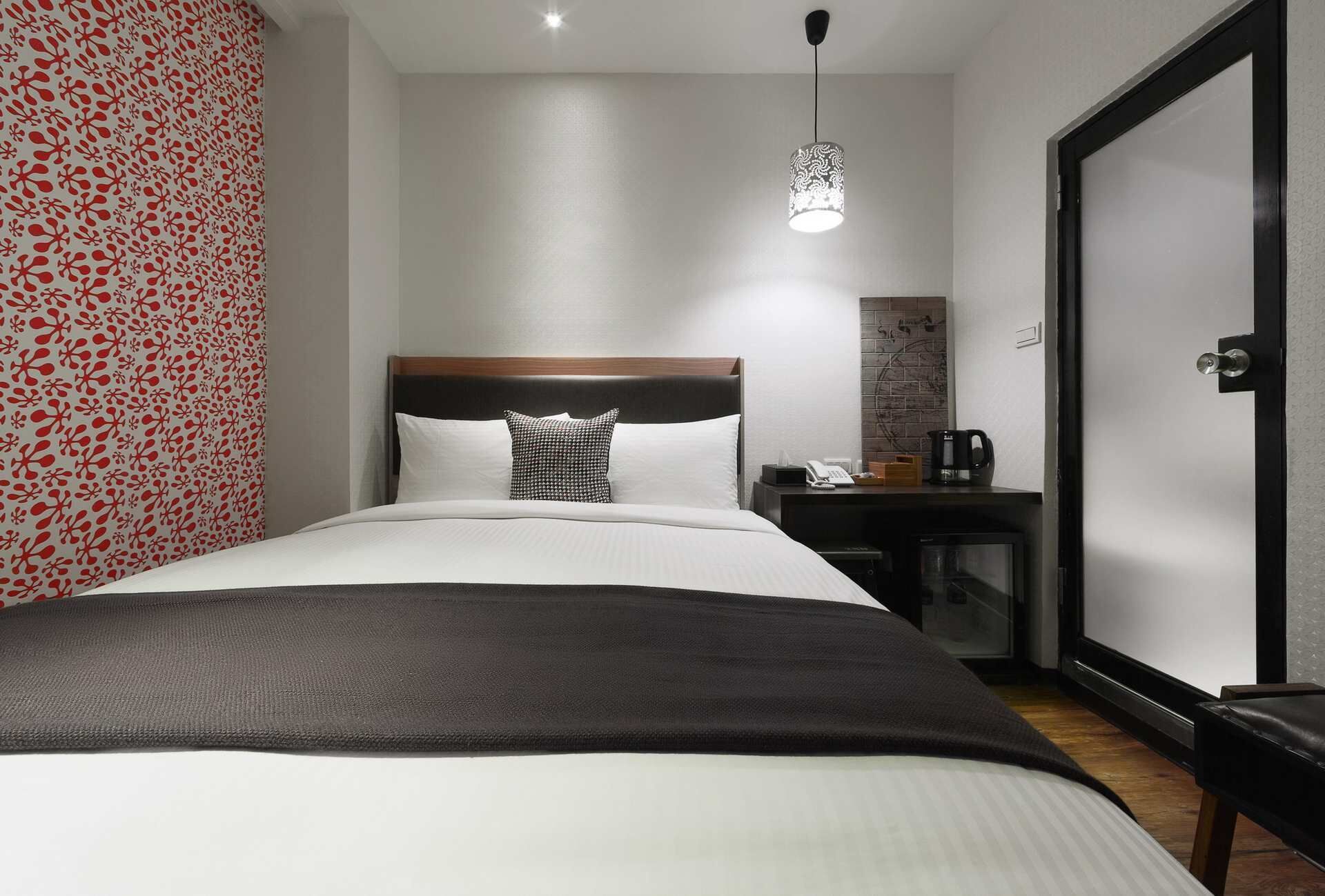 Habitación de hotel con cama, mesita de noche con teléfono y cafetera, pared con diseño rojo, y puerta.