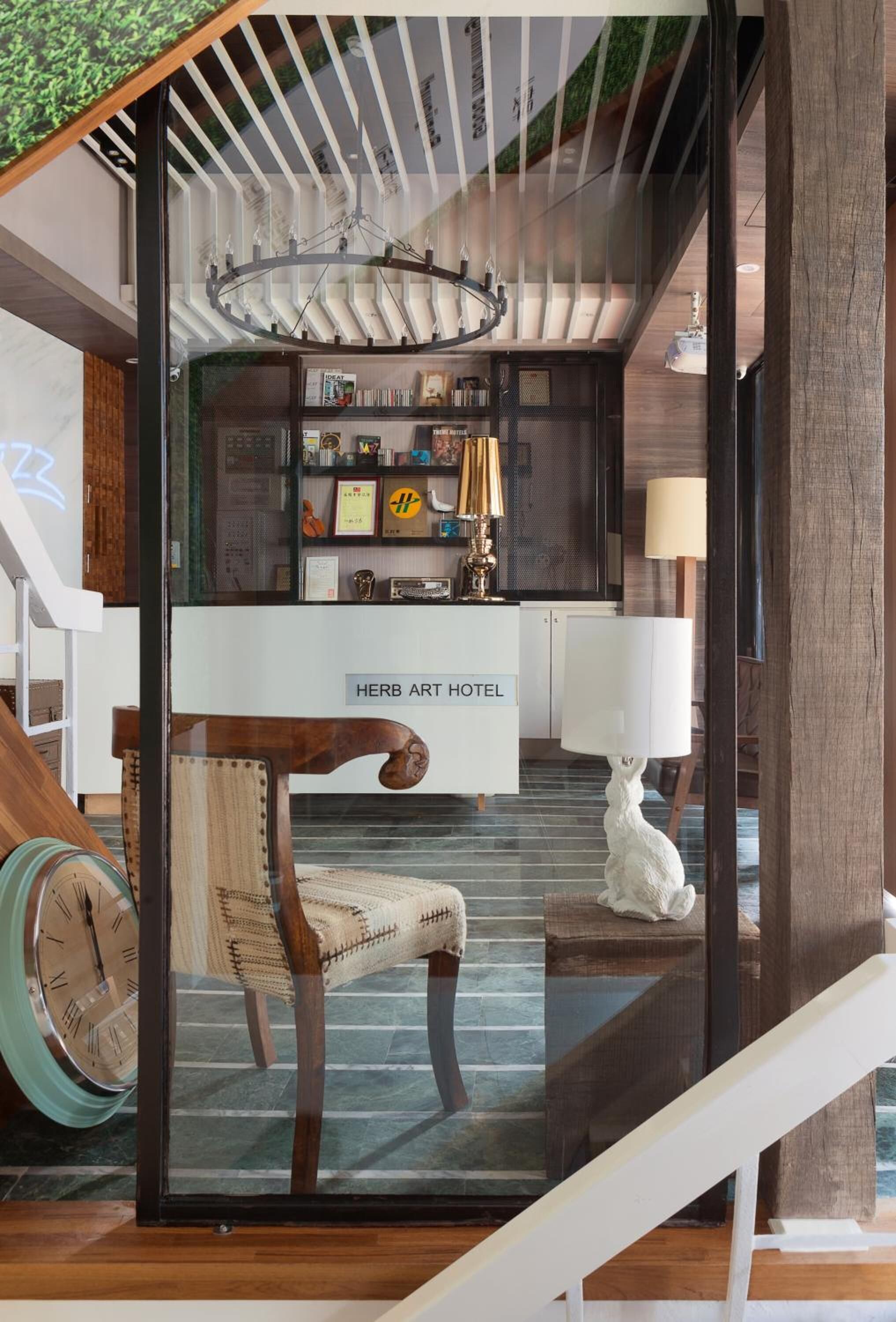 A modern interior with a wooden staircase, a reception desk labeled "HERB ART HOTEL," and a chandelier.