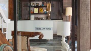 A modern interior with a wooden staircase, a reception desk labeled "HERB ART HOTEL," and a chandelier.