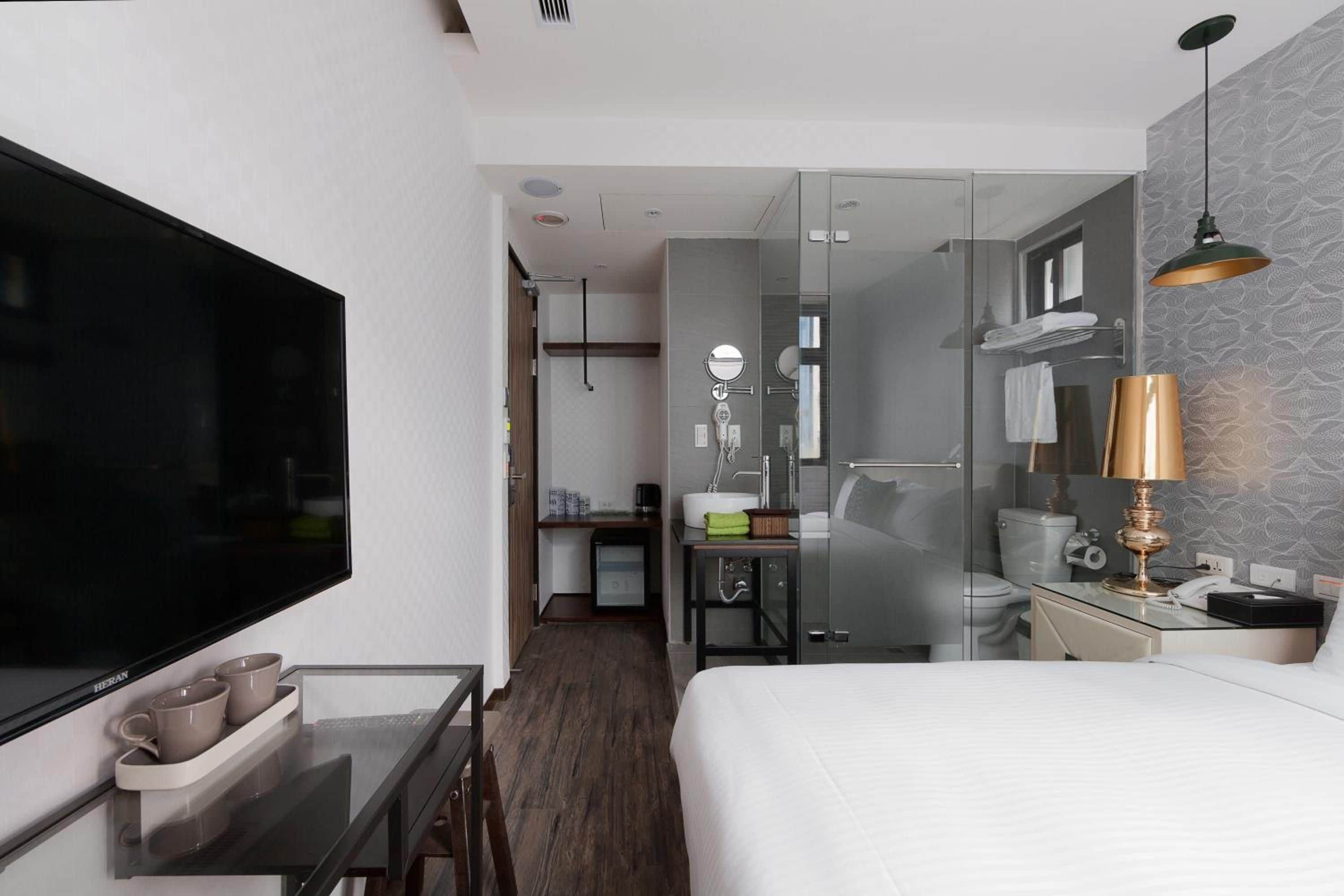 Quarto de hotel com cama, secretária com televisão de tela plana, box de vidro para o chuveiro e pequena cozinha.