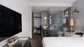 Quarto de hotel com cama, secretária com televisão de tela plana, box de vidro para o chuveiro e pequena cozinha.