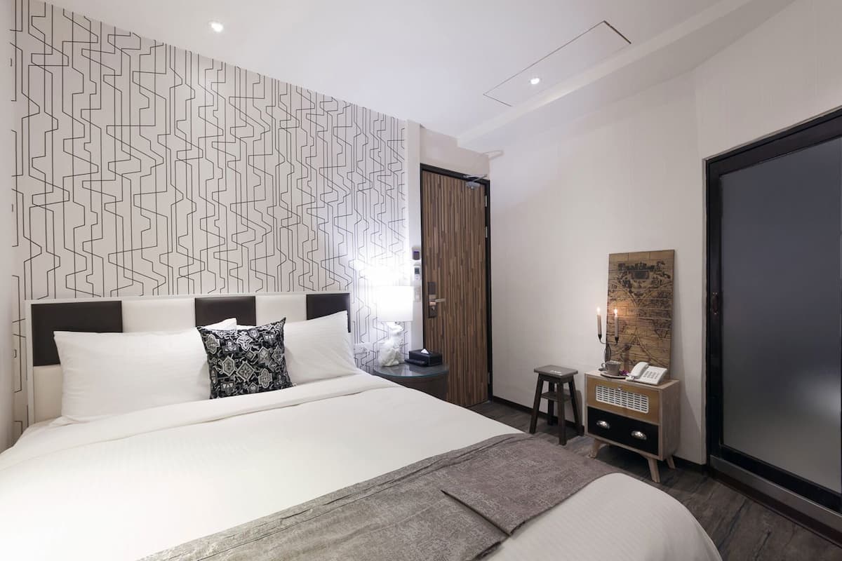 Una habitación de hotel moderna con una cama grande, un papel tapiz decorado, una puerta de madera, una mesita con un teléfono y un espejo amplio.