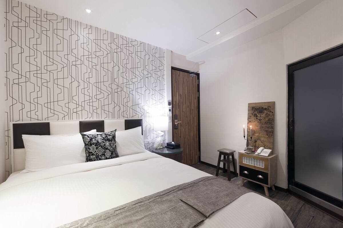 Habitación de hotel moderna con una cama grande, un papel tapiz estampado, una puerta de madera, una mesita de noche con lámpara y un espejo grande.