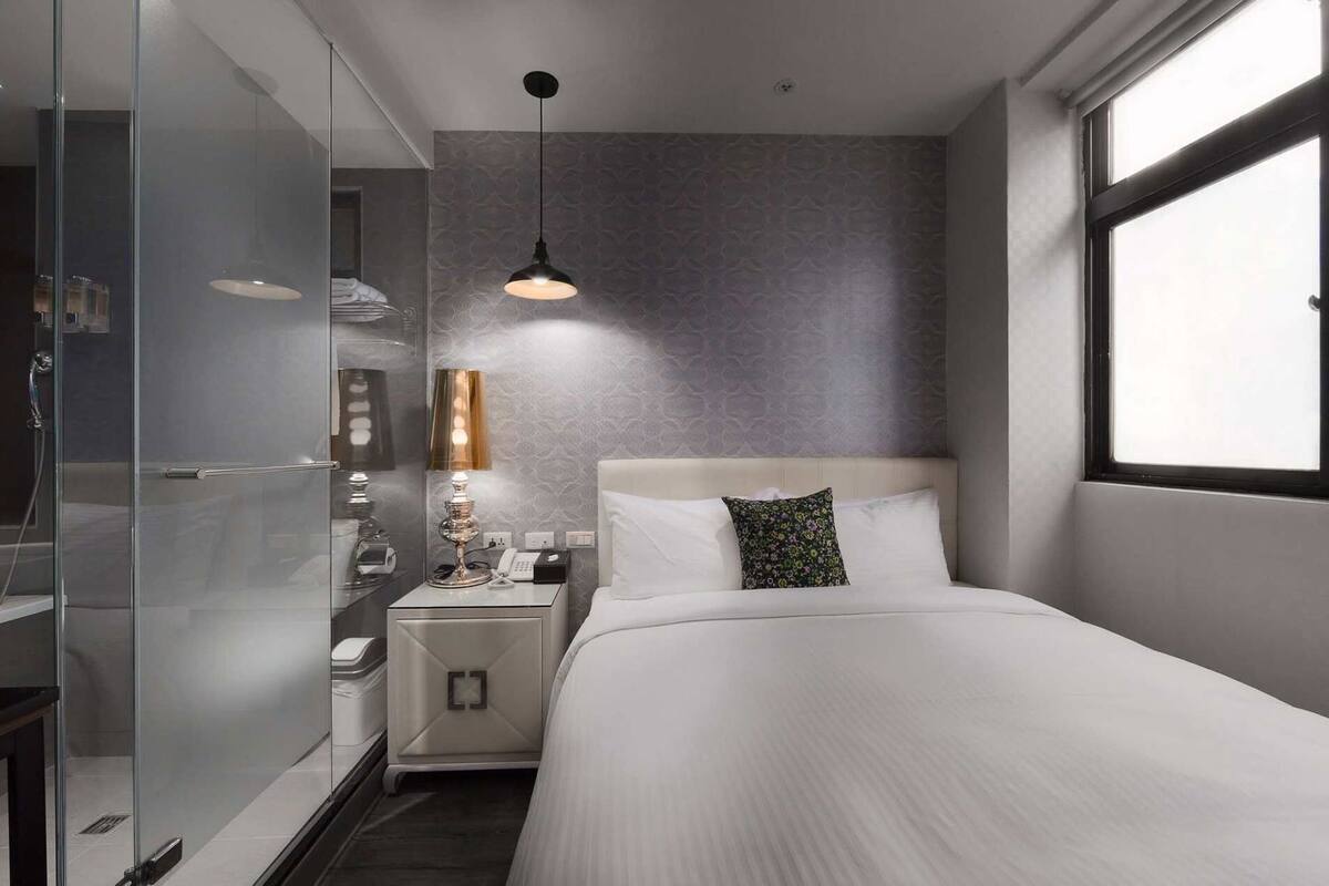 Una habitación de hotel moderna con cama, mesita de noche, lámpara y ventana.