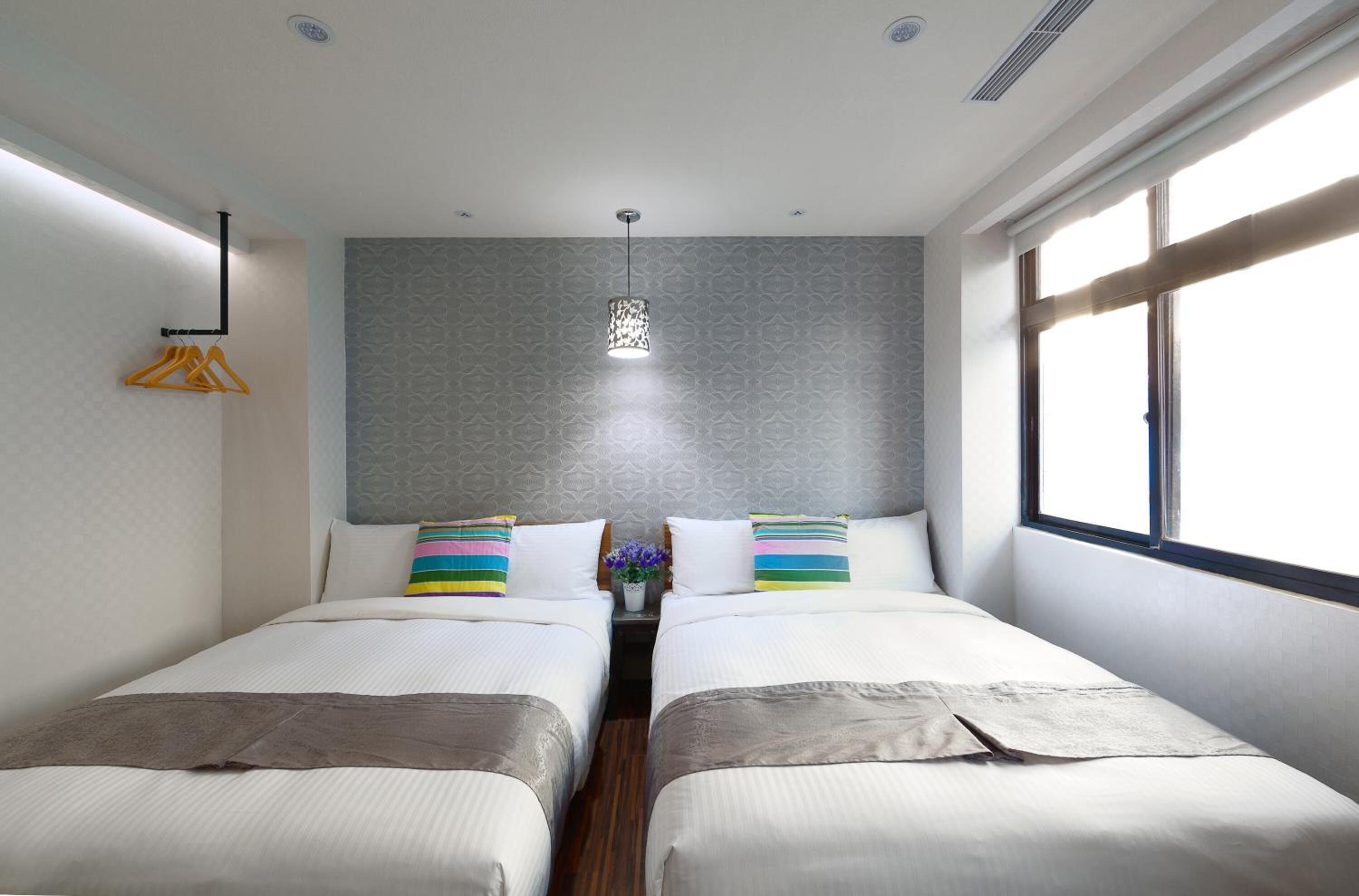 Ein modernes Hotelzimmer mit zwei Betten, einer Pendelleuchte und einem Fenster mit Jalousien.