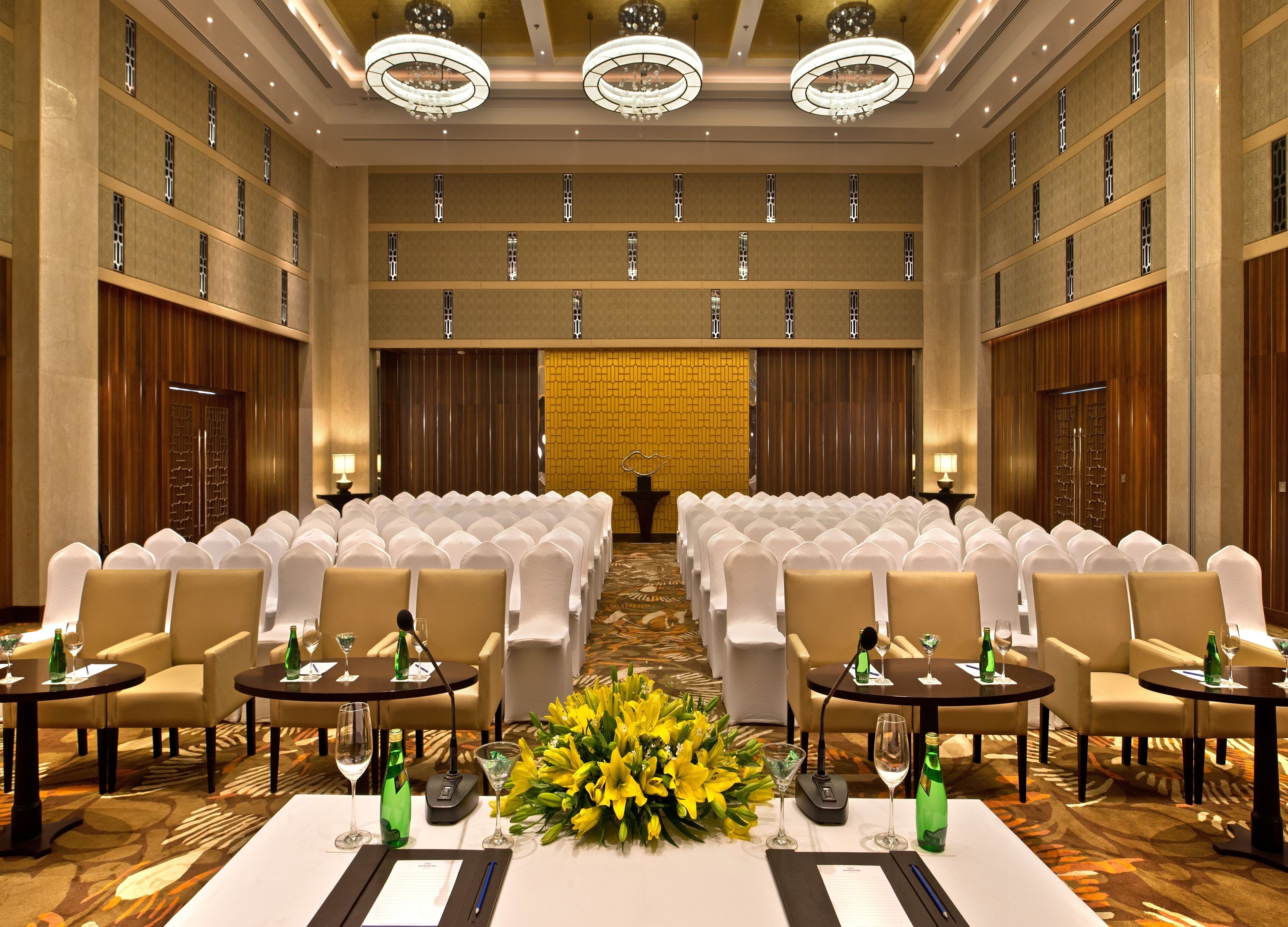 A conference room with rows of white chairs, tables with microphones, and a floral centerpiece.
