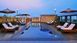 Un área de piscina en la azotea con sillones de descanso y sombrillas, un bar y vistas a un paisaje urbano.