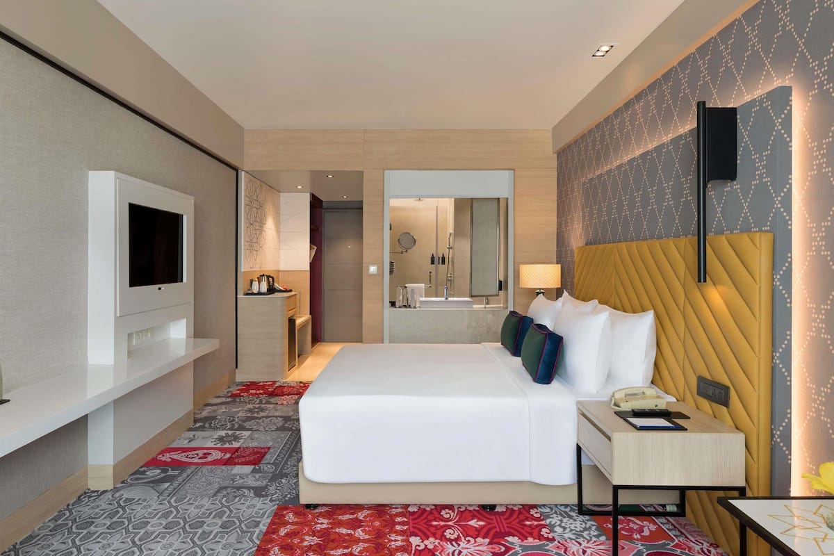 A hotel room with a bed, a TV, a desk with a lamp, and a bathroom visible through an open door.