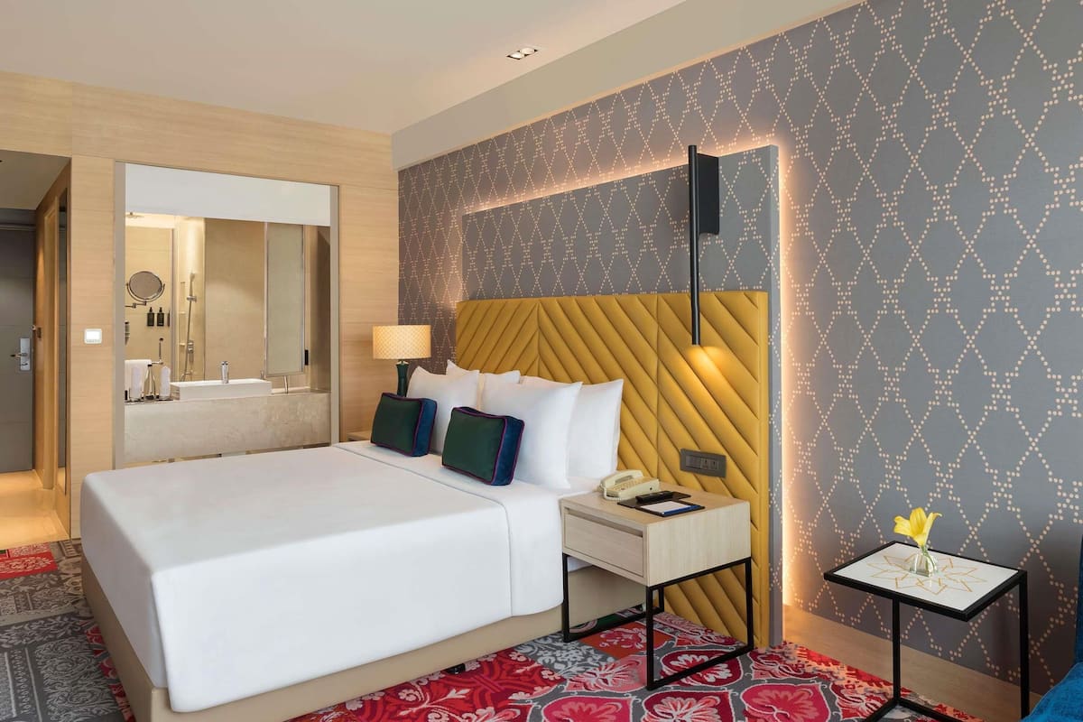 A hotel room with a bed, bedside table, lamp, and a small table with a vase.