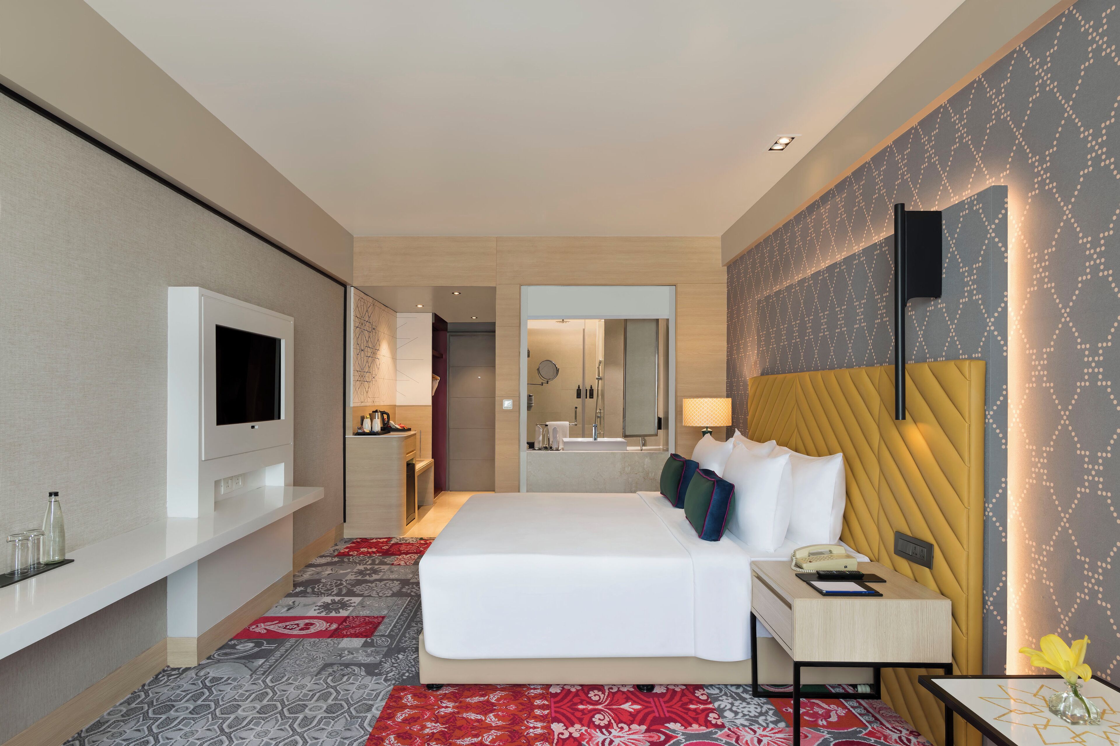 A modern hotel room with a large bed, a flat-screen TV, a bedside table with a lamp, and a mirror above the bed.