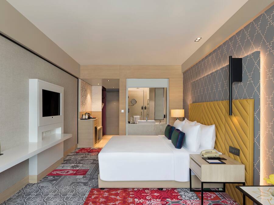 A modern hotel room with a large bed, a flat-screen TV, a bedside table with a lamp, and a mirror above the bed.