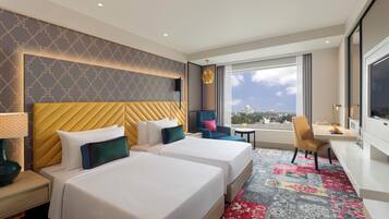 Habitación de hotel con dos camas, un escritorio, una silla, un televisor y un ventanal con vistas a la ciudad.