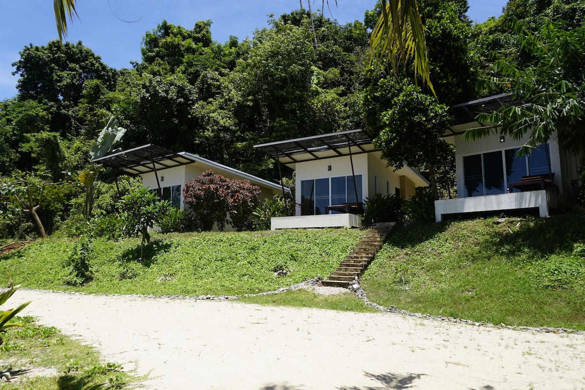 A modern house with a covered patio, surrounded by lush greenery and a sandy path leading to the entrance.