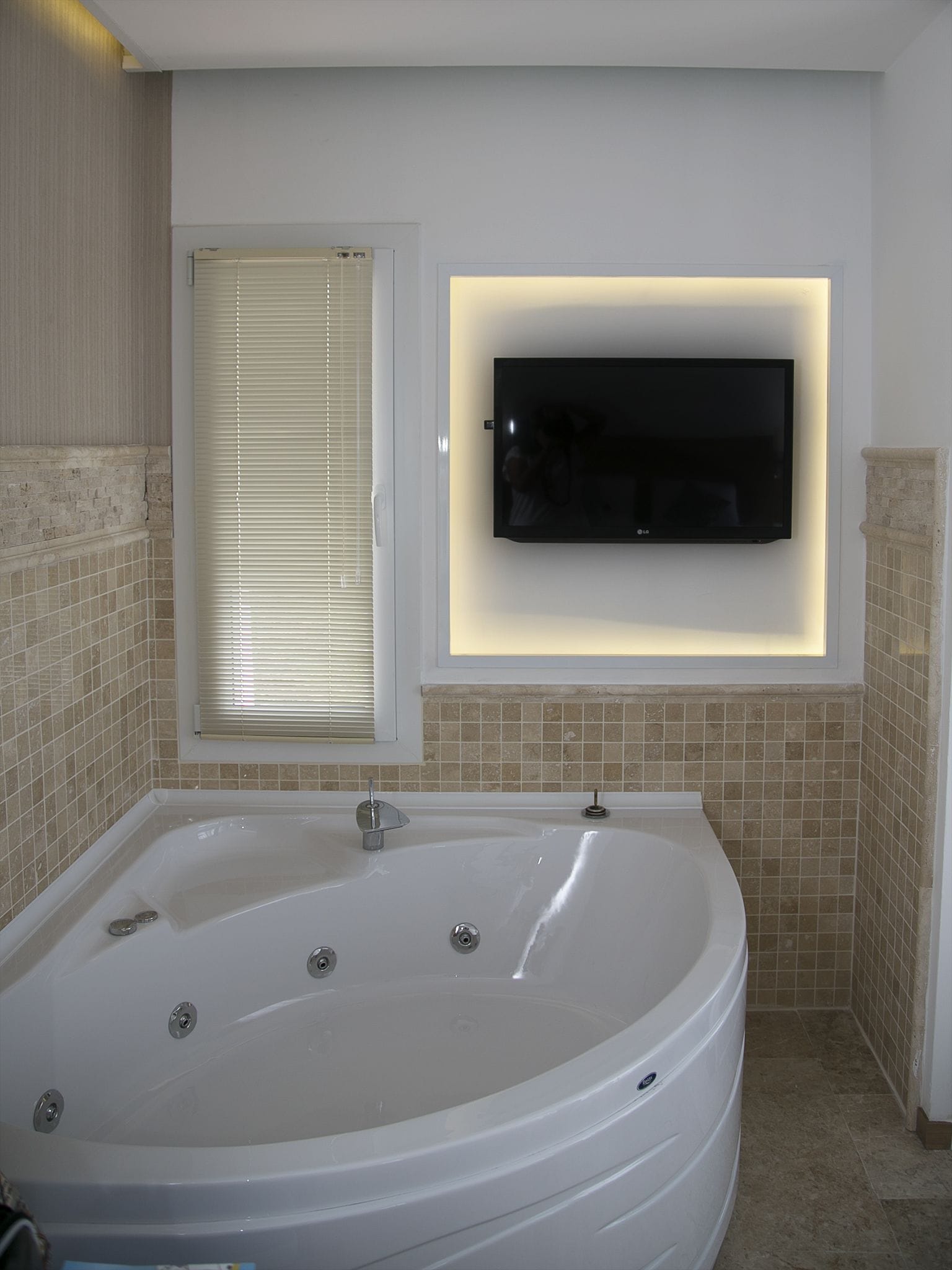 A modern bathroom with a large bathtub, a wall-mounted TV, and a window with blinds.