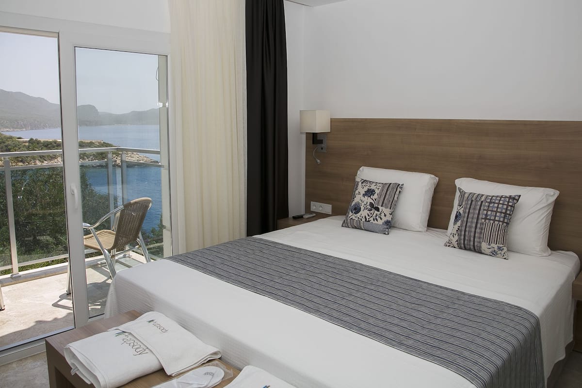 A hotel room with a large bed, a nightstand with a lamp, and a view of the sea through a sliding glass door.