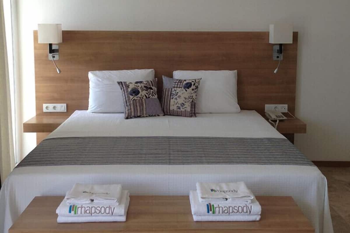 A hotel room with a wooden headboard, two white pillows, a gray blanket, and two bedside tables with folded towels.