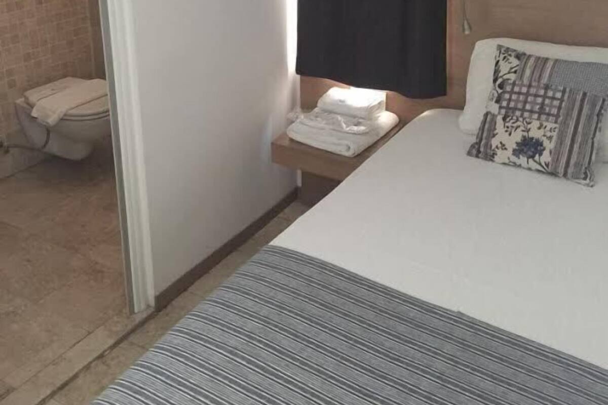 A hotel room with a bed, a bedside table with a lamp, a bathroom with a toilet and shower, and a window with a curtain.