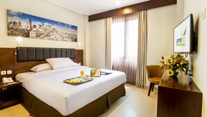 A hotel room with a large bed, a cityscape painting, a desk with a computer, and a window with curtains.