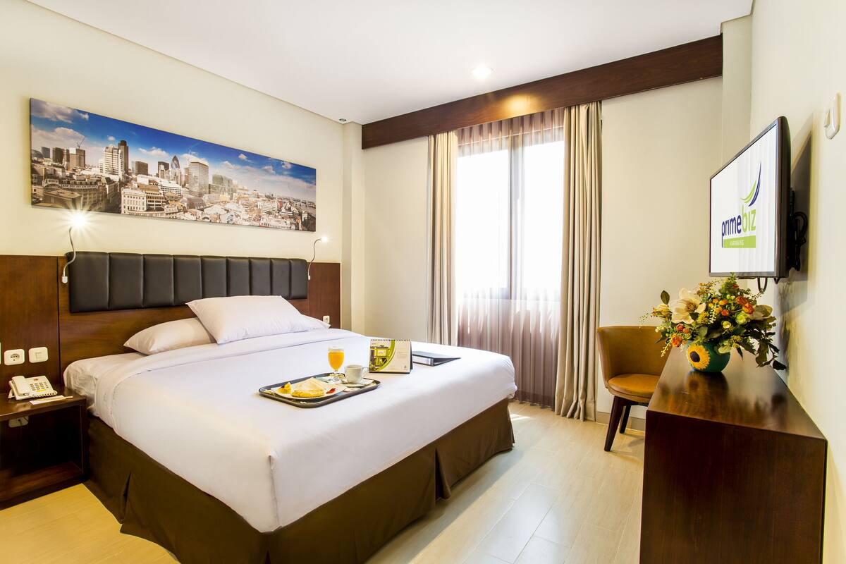Een hotelkamer met een groot bed, een stadsgezicht schilderij, een bureau met een computer en een raam met gordijnen.