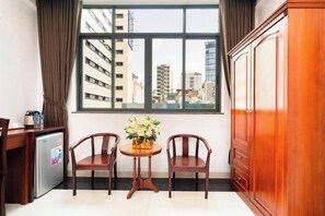 A hotel room with a large window, two wooden chairs, a small round table with a vase of flowers, a desk with a kettle and water bottles, and a wooden wardrobe. - Hoang Hoang Hotel (Ho Chi Minh City)