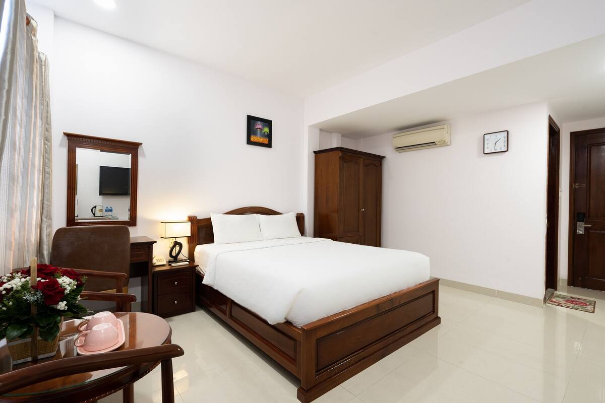 A neatly arranged bedroom with a bed, a chair, a desk, a mirror, a clock, and an air conditioning unit.