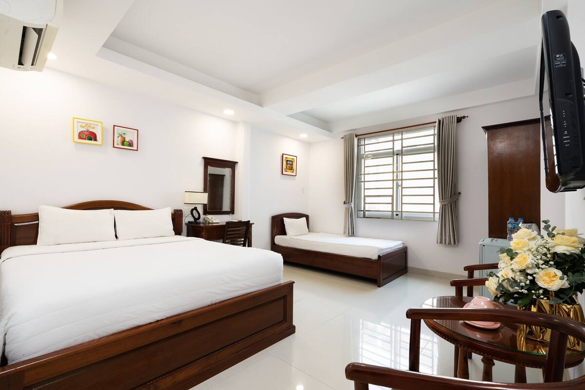 A hotel room with a large bed, a smaller bed, a desk with a chair, a TV mounted on the wall, and a window with curtains.