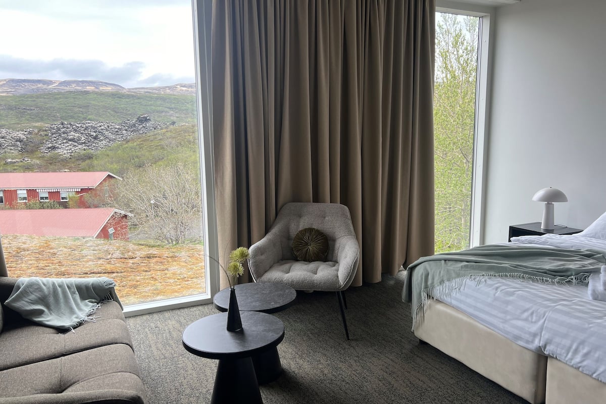 Habitación de hotel con un ventanal grande, una cama, una silla, una mesita y una lámpara.