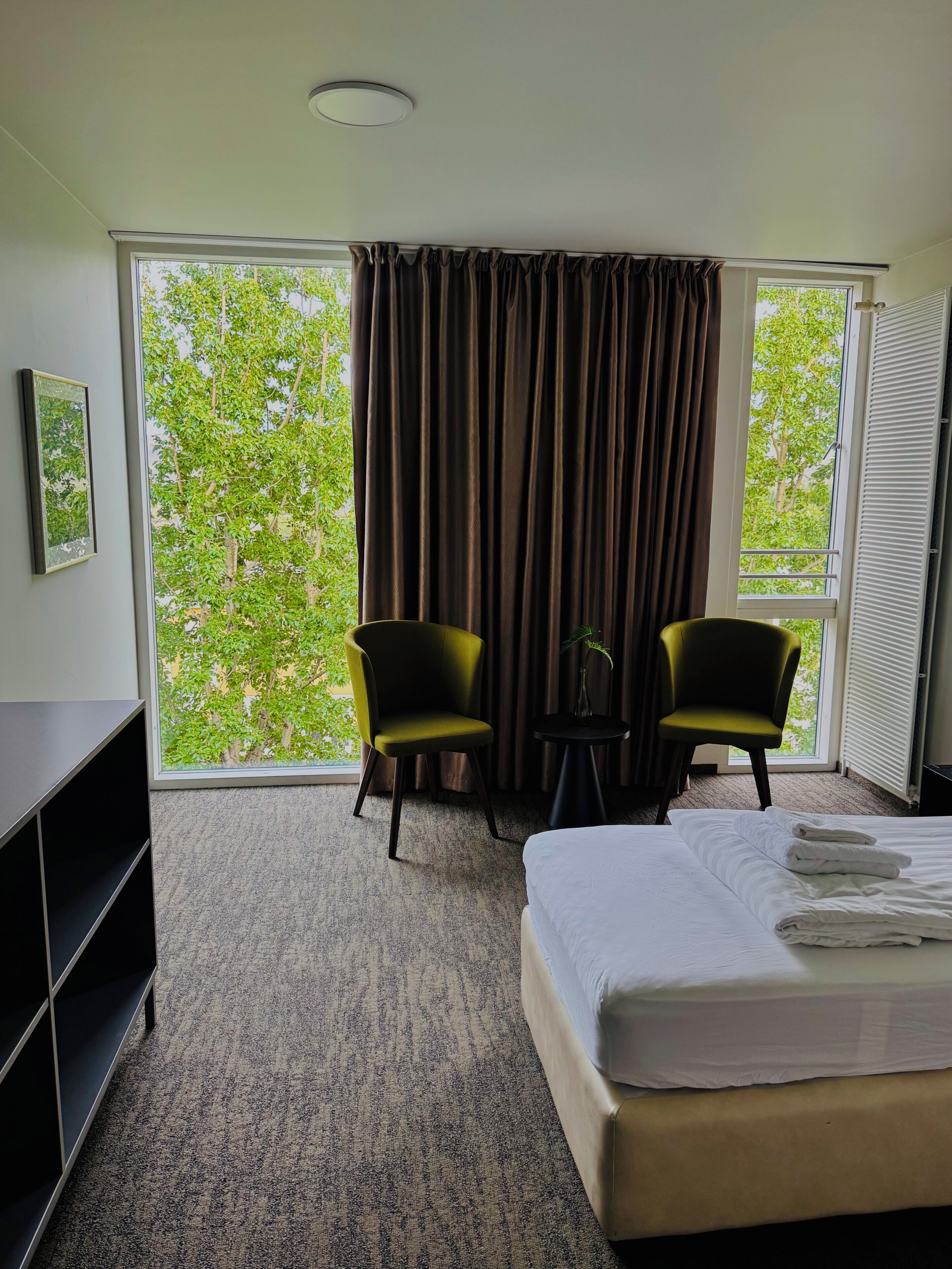 A hotel room with a bed, two chairs, a small table, a window with curtains, and a picture on the wall.