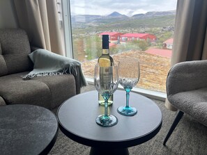 A room with a round table, two chairs, a bottle of wine, and two wine glasses. A window offers a view of buildings and mountains. - Hotel Bifrost (Borgarbyggd)