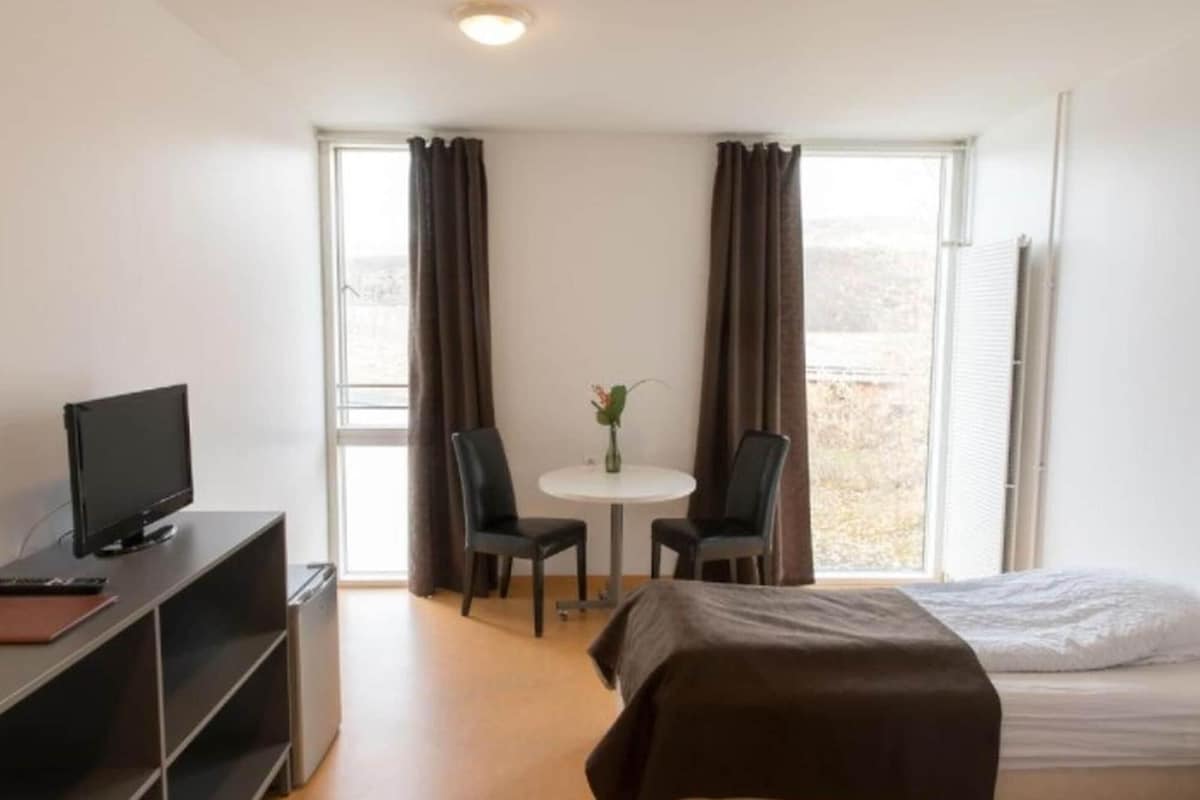 A hotel room with a bed, a desk with a computer, a small table with two chairs, and a window with curtains.