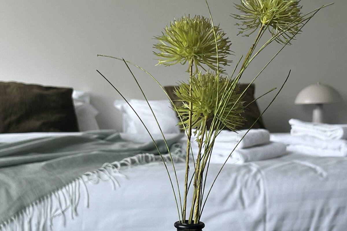 Una cama con ropa blanca y una manta verde, un jarrón negro con plantas verdes y una lámpara de noche.