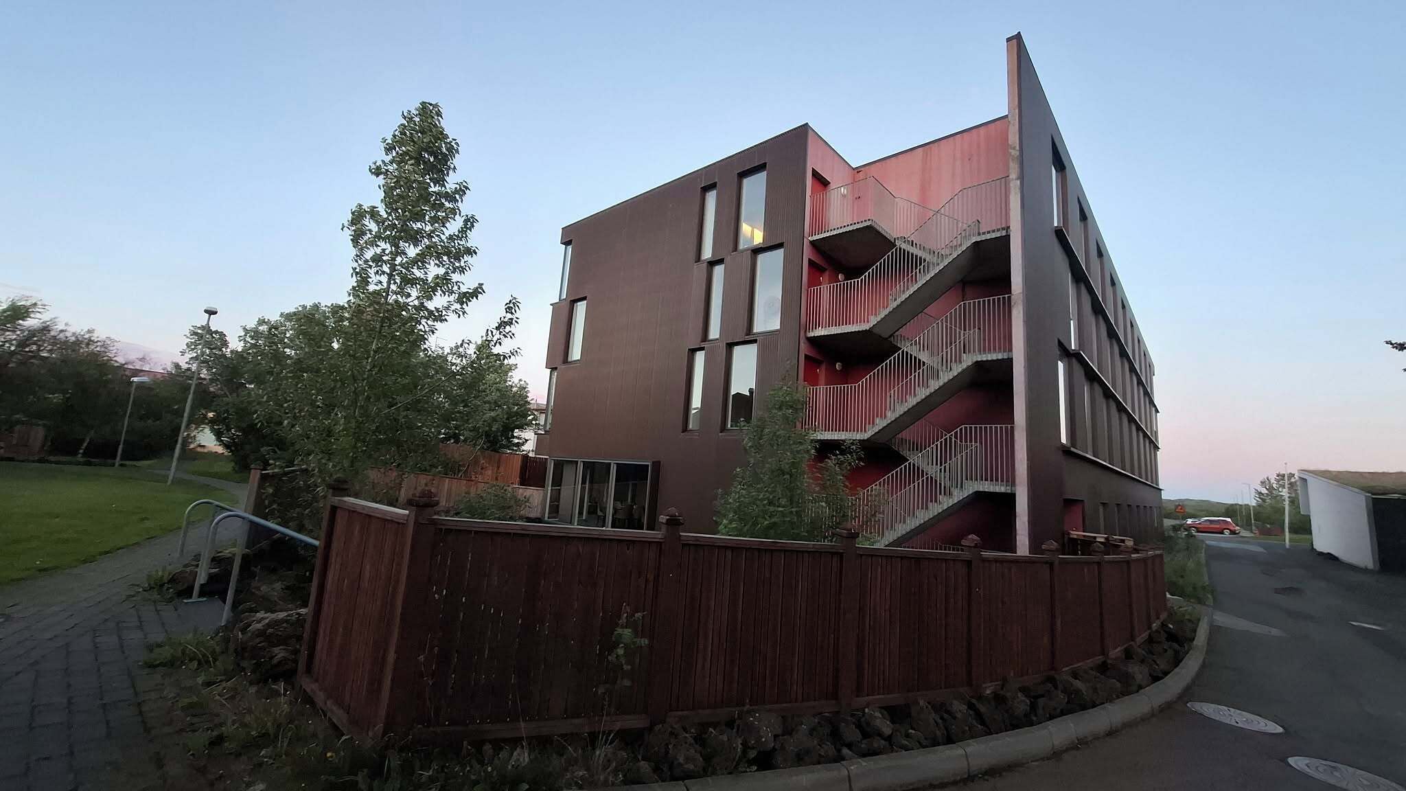 A multi-story building with balconies, a wooden fence, and a paved driveway.