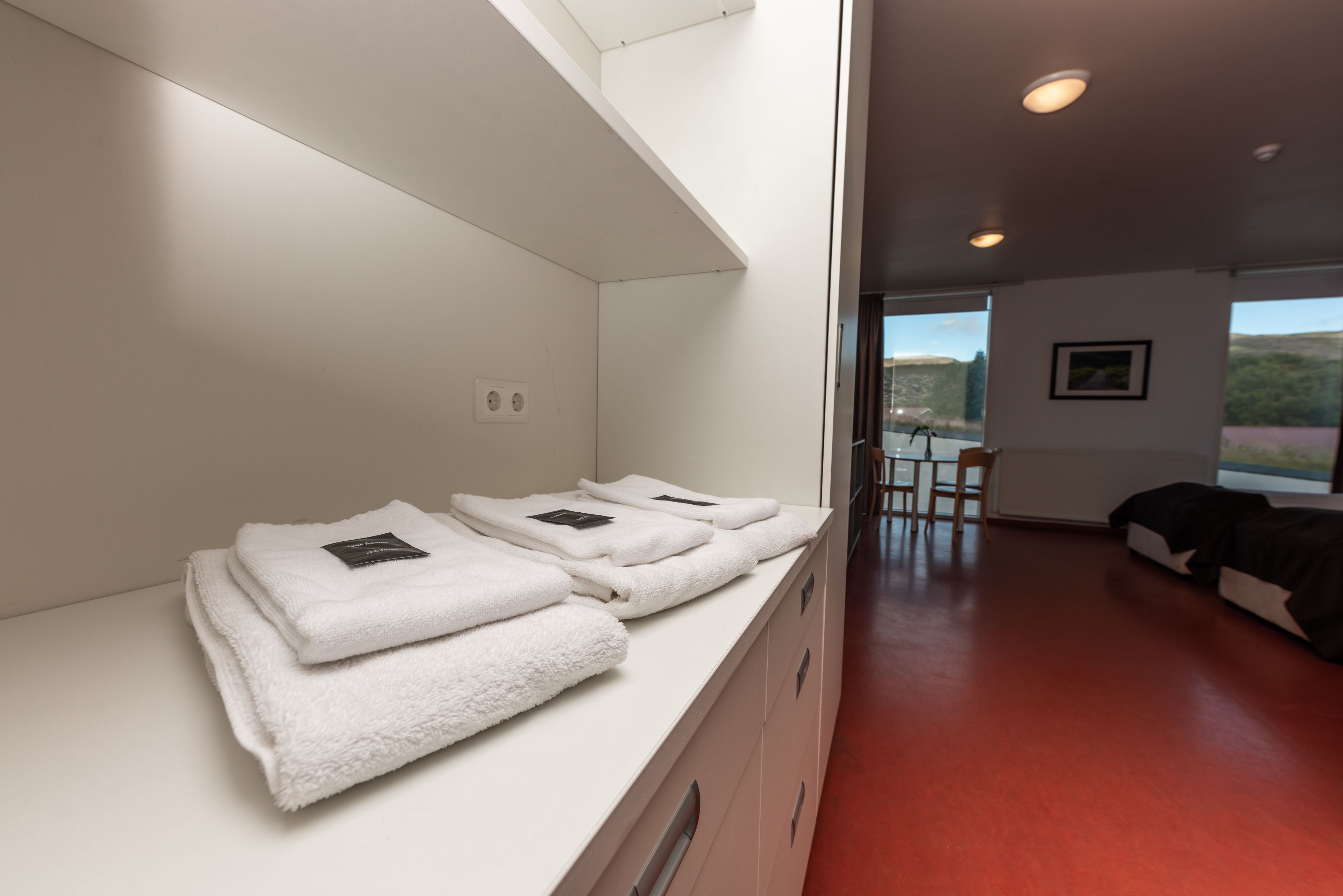 Ein Hotelzimmer mit einer weißen Theke, auf der gefaltete Handtücher liegen, einem Holzstuhl, einer schwarzen Couch und Blick ins Freie durch ein großes Fenster.