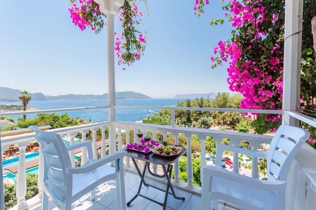 A balcony with white chairs, a table with fruit and drinks, and a view of the sea and mountains.
