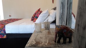 A bed with white bedding and colorful pillows, a small table with jars and a decorative elephant figure, and a window with a view of the sky. - Anara Villa (Unawatuna)
