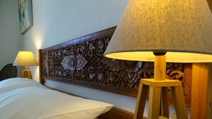 Une chambre d’hôtel avec un lit soigneusement fait, une tête de lit en bois ornée de motifs complexes et une lampe posée sur un piédestal en bois.