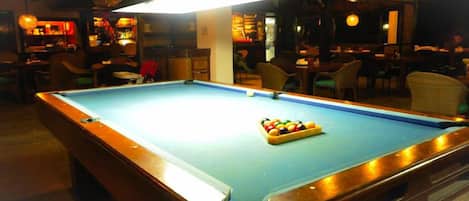 A pool table with a light fixture above it, surrounded by chairs and tables in a dimly lit room.