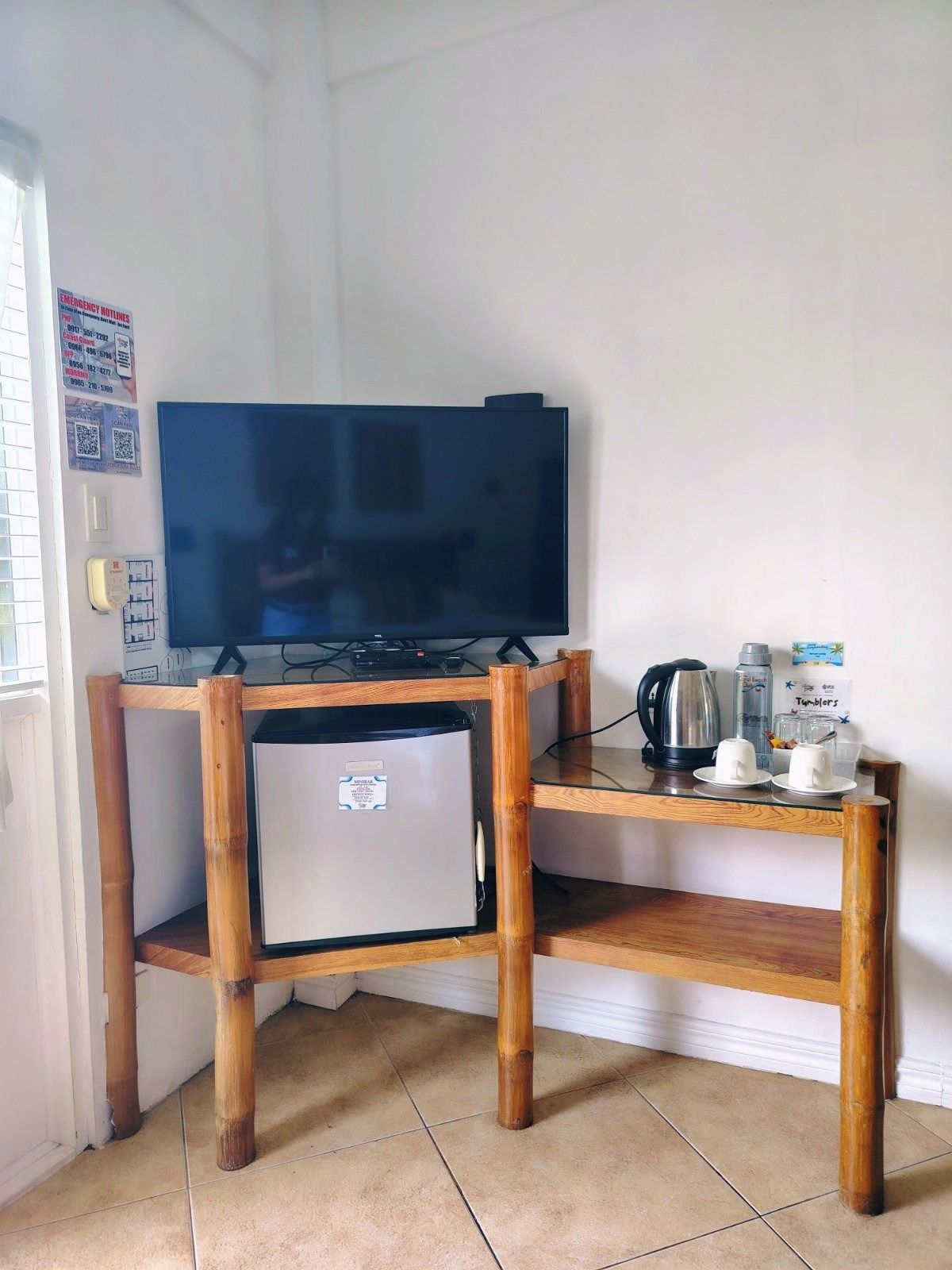 A compact room with a television on a wooden stand, a small refrigerator, a coffee maker, and a kettle.