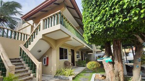 A two-story building with a staircase leading to the entrance, a small porch area, and a garden with a tree and shrubs. - The Coral Beach Club (Lian)