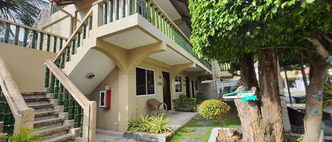A two-story building with a staircase leading to the entrance, a small porch area, and a garden with a tree and shrubs.