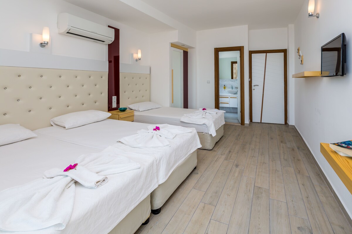Quarto de hotel com três camas, uma televisão e um banheiro visível através de uma porta aberta.