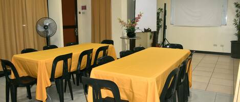 Un salón de conferencias con dos mesas cubiertas con manteles amarillos, sillas negras, una pizarra, un reloj y un ventilador.